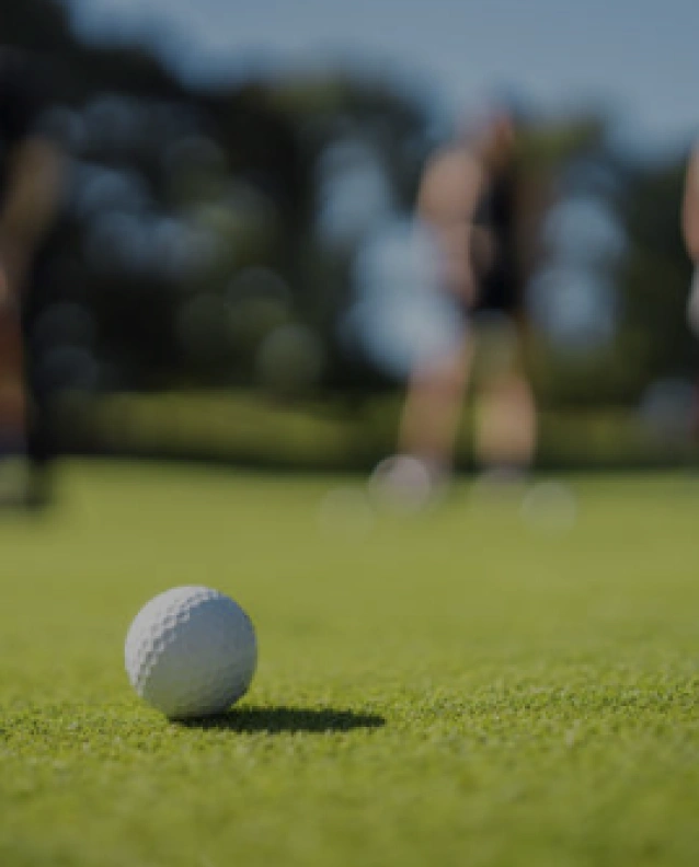 Close-up of golf ball on green grass with blurred players preparing in the background