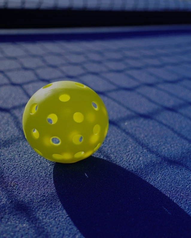 Bright yellow pickleball resting on a blue court with dramatic shadow from nearby net