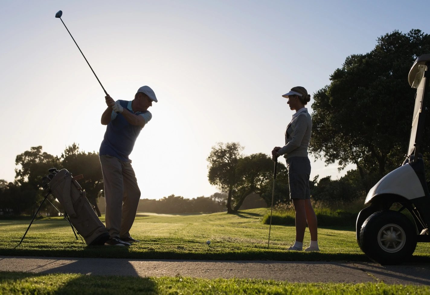 Man swinging golf club while woman watches beside golf cart at scenic green course