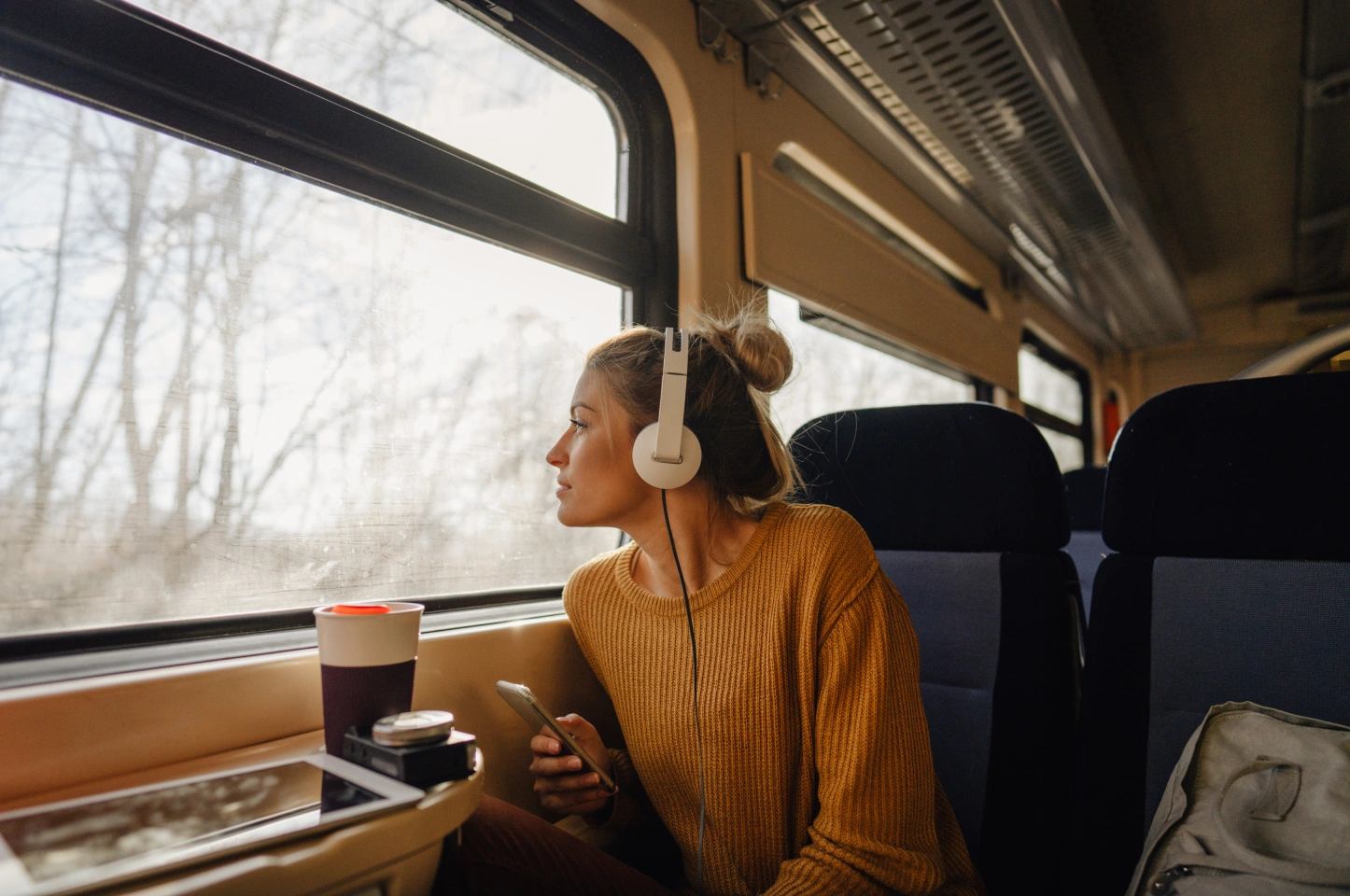Woman with headphones looking out train window while holding phone during scenic ride